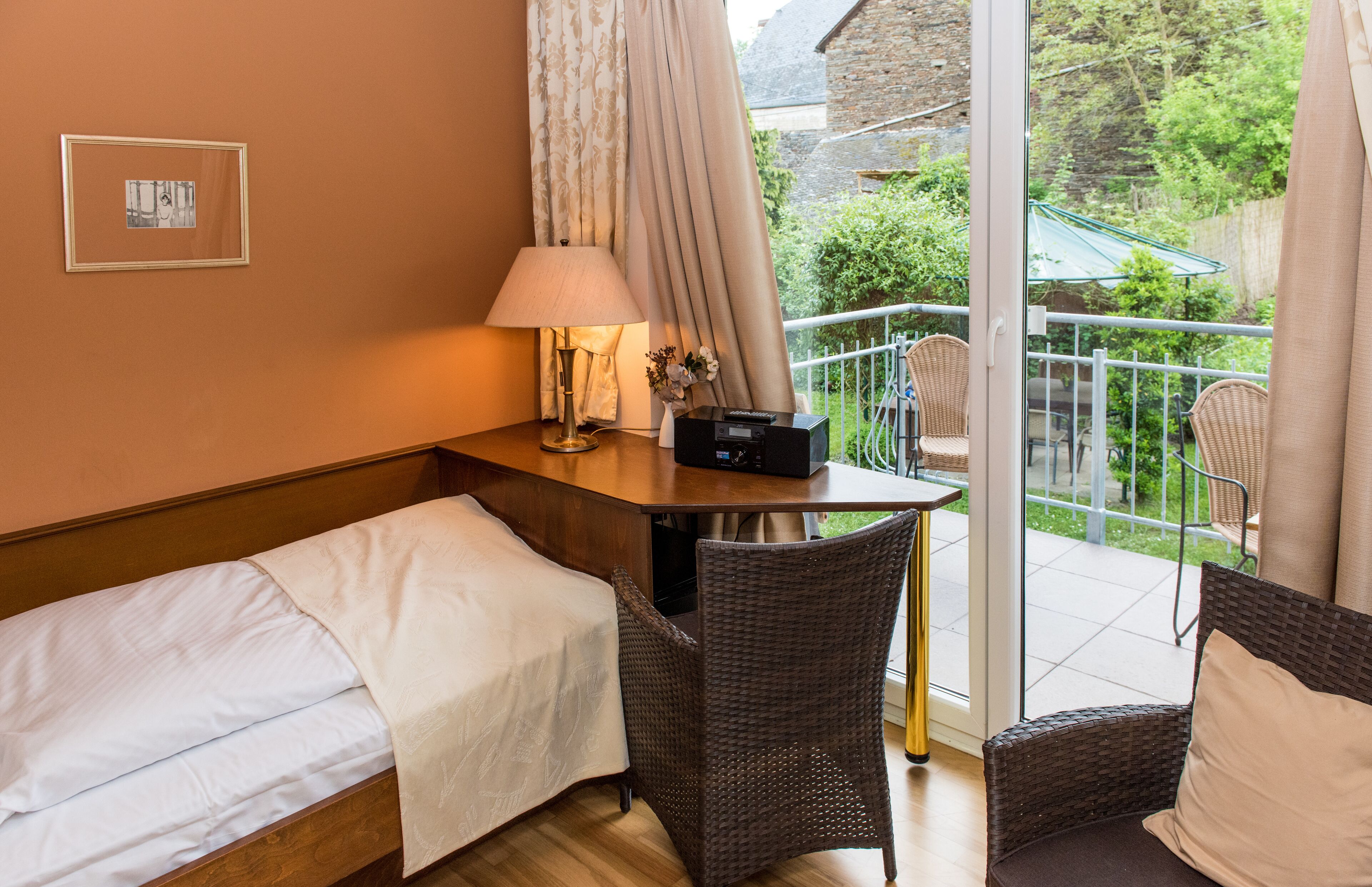 Habitación de hotel con cama, escritorio con lámpara, silla y balcón con mobiliario de mimbre y vistas a la vegetación.