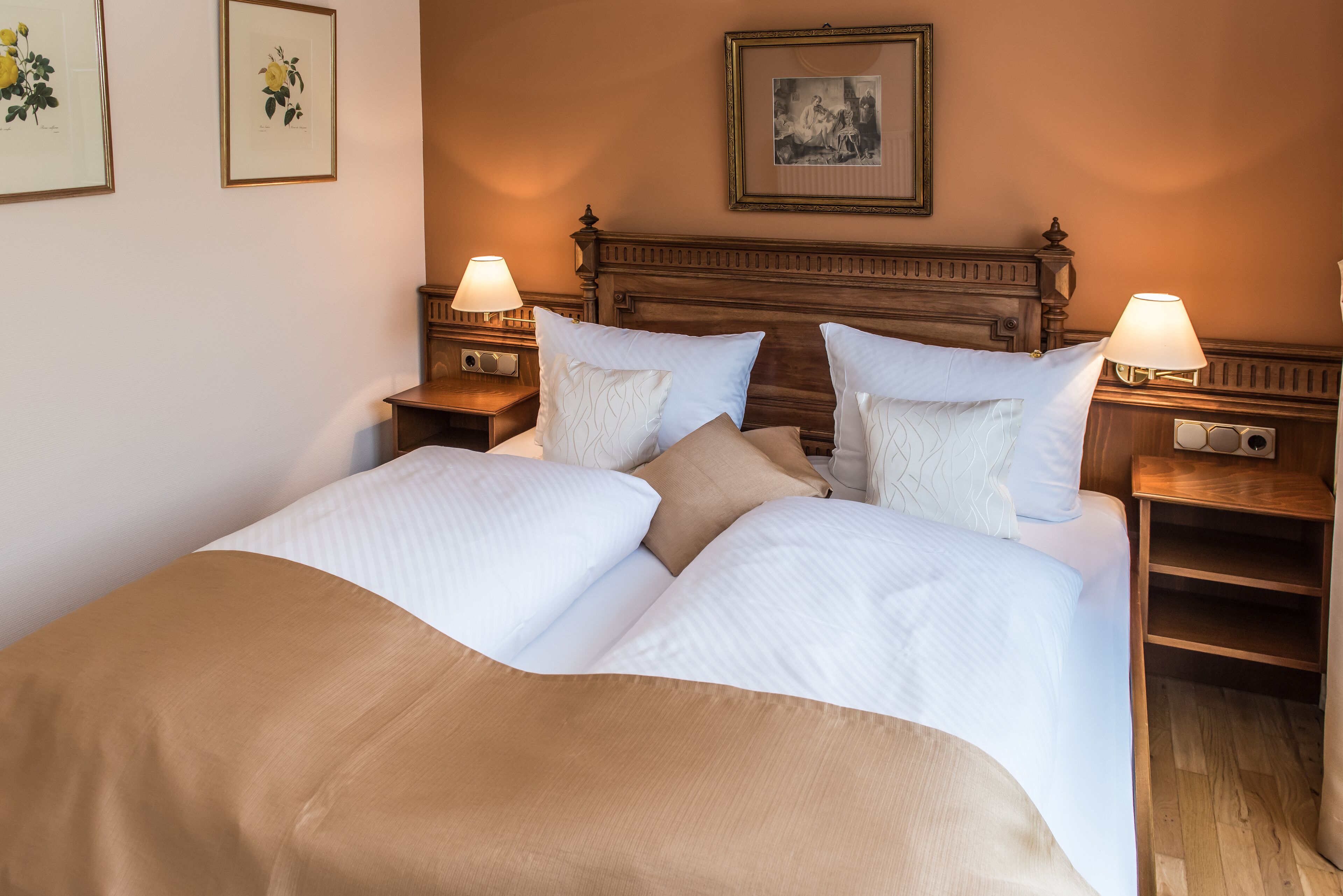Una cama doble con edredón color crema y blanco, dos mesitas de noche con lámparas, un cabecero de madera e impresiones botánicas enmarcadas en la pared.