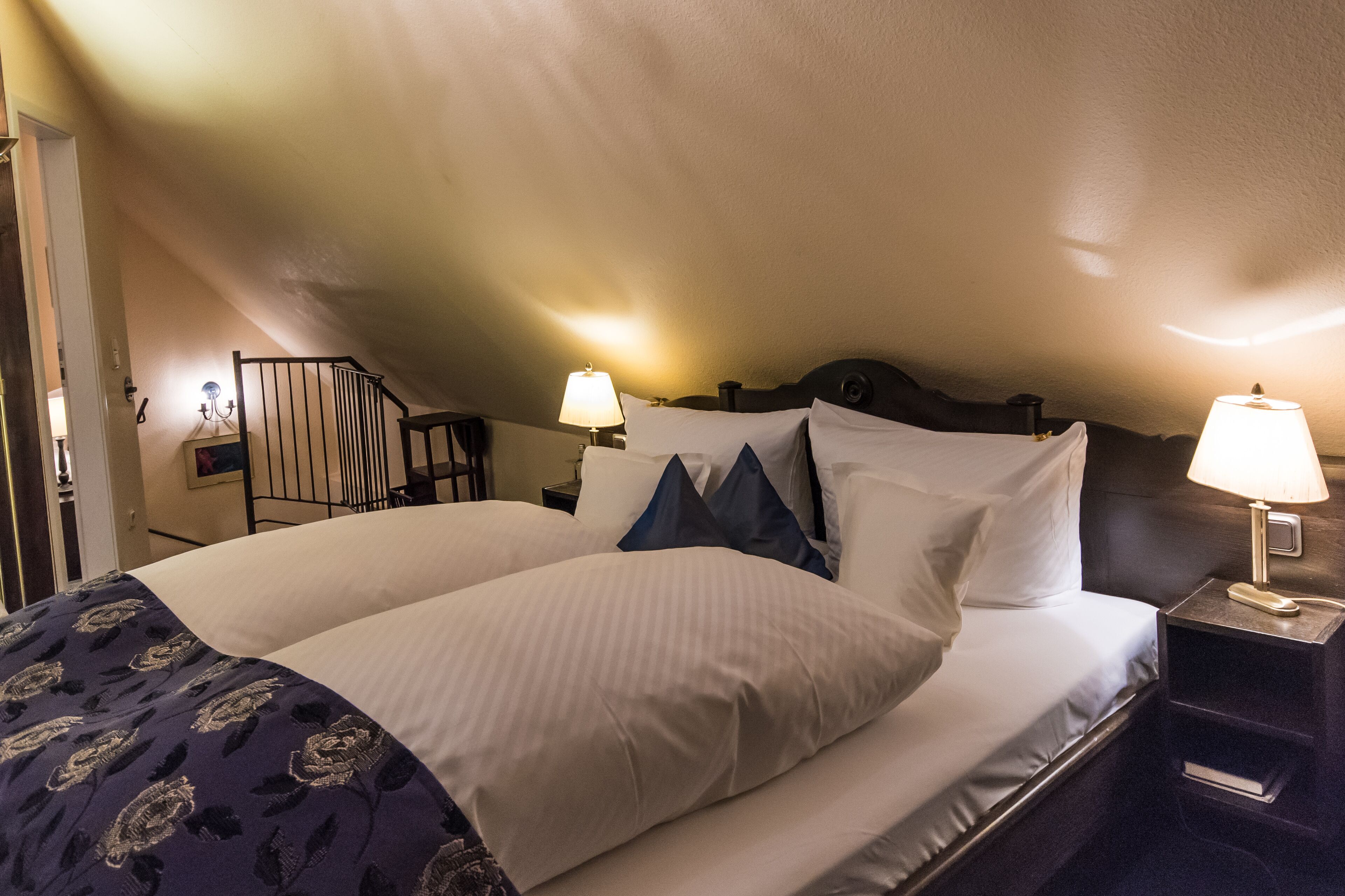 Ein ordentlich bezogenes Bett mit weißen und blauen Kissen, einer gemusterten Tagesdecke und zwei Nachttischlampen.