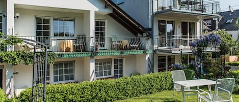 Una casa de dos pisos con balcón, un pequeño jardín y una mesa blanca con sillas.