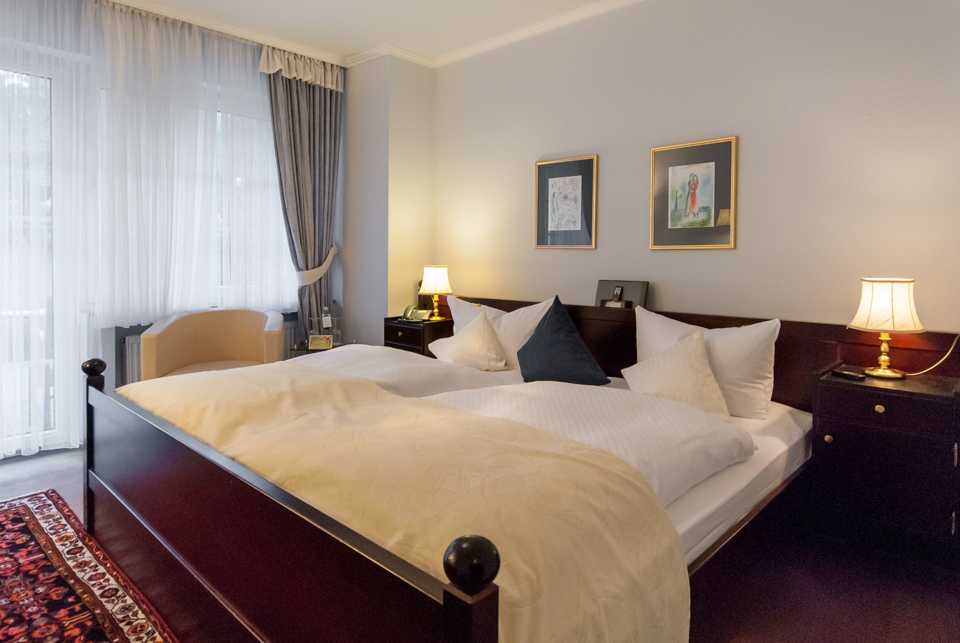 A hotel room with a large bed, two bedside tables, a nightstand, a window with curtains, and two framed pictures on the wall.