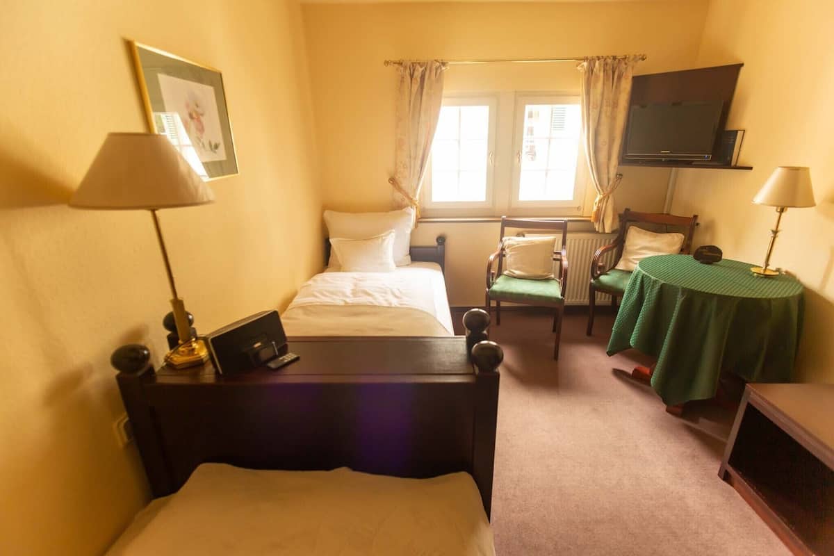 Hotellihuone, jossa on kaksi erillistä sänkyä, pieni pöytä vihreällä pöytäliinalla, tuoli, seinälle asennettu televisio ja ikkuna, jossa on verhot.