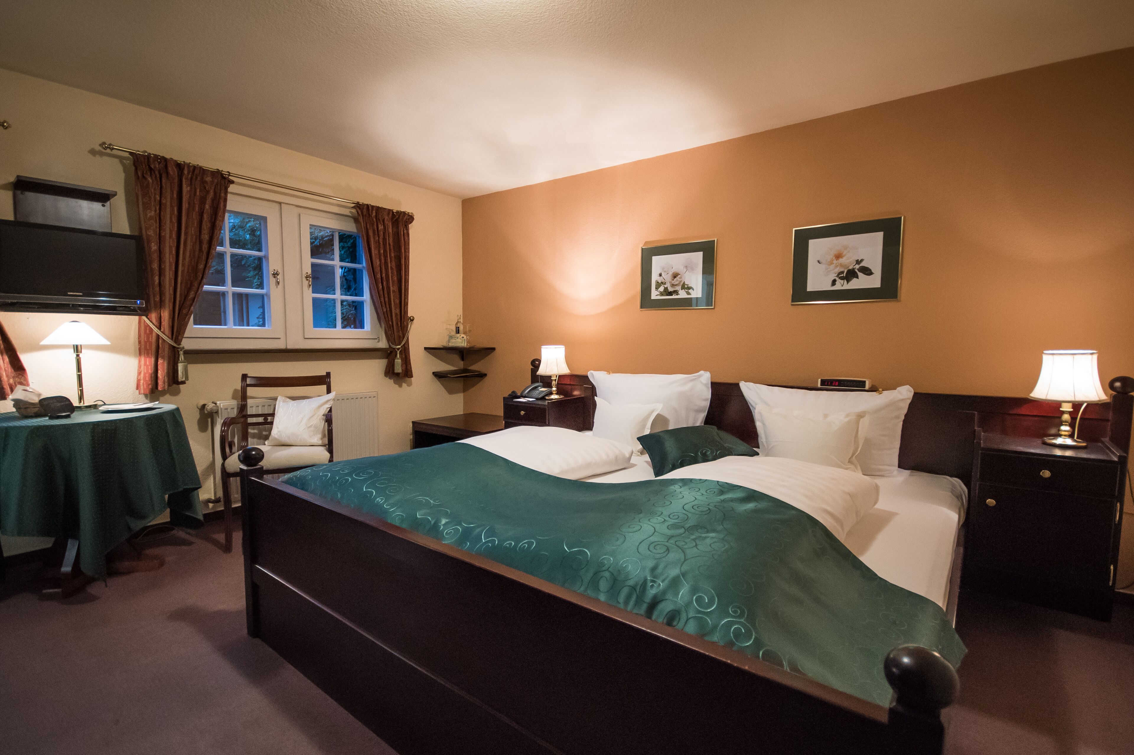 A hotel room with a bed, bedside tables, a desk, a chair, a TV, and two framed pictures on the wall.