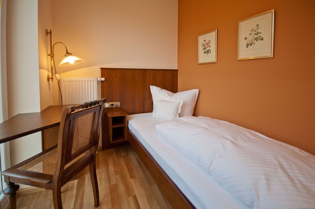 A small, neatly arranged bedroom with a wooden desk, a chair, a bed with white bedding, and two framed botanical prints on the wall.
