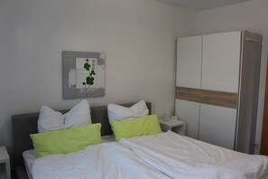 Ein ordentlich bezogenes Bett mit weißen und grünen Kissen, ein Nachttisch mit Lampe sowie ein Kleiderschrank mit Schiebetüren.