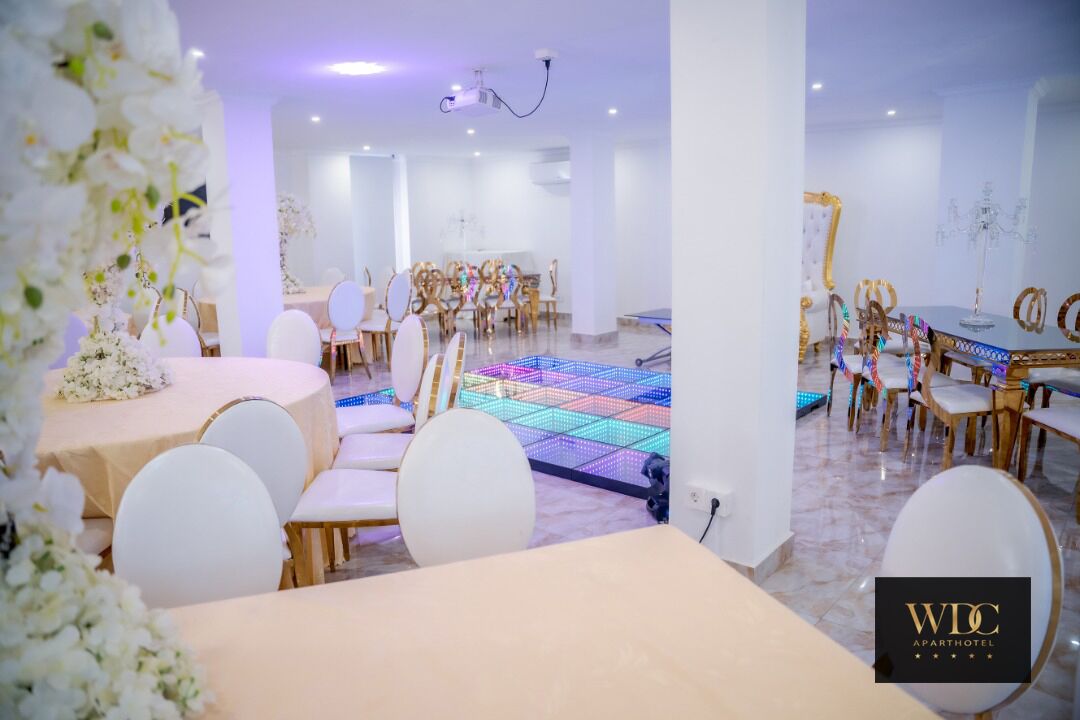 A spacious room with multiple round tables and white chairs, a colorful LED dance floor, and a chandelier.