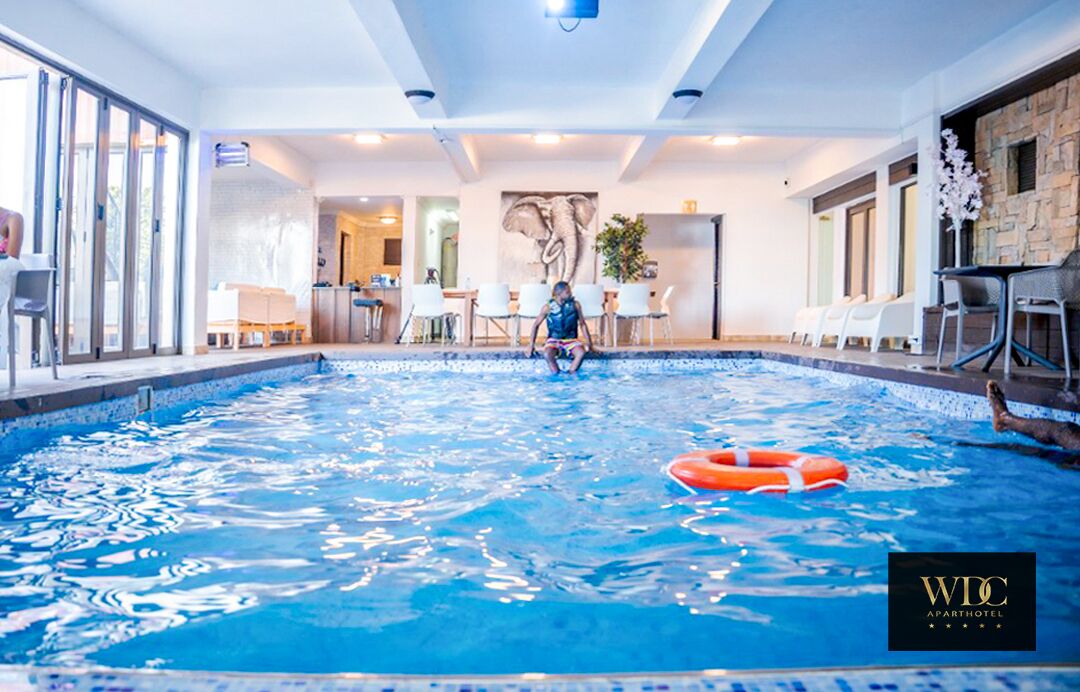 Piscina interior com boia salva-vidas, uma pessoa sentada à beira da piscina e uma área de recepção com cadeiras e mesa.