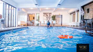 Piscina interior com boia salva-vidas, uma pessoa sentada à beira da piscina e uma área de recepção com cadeiras e mesa.
