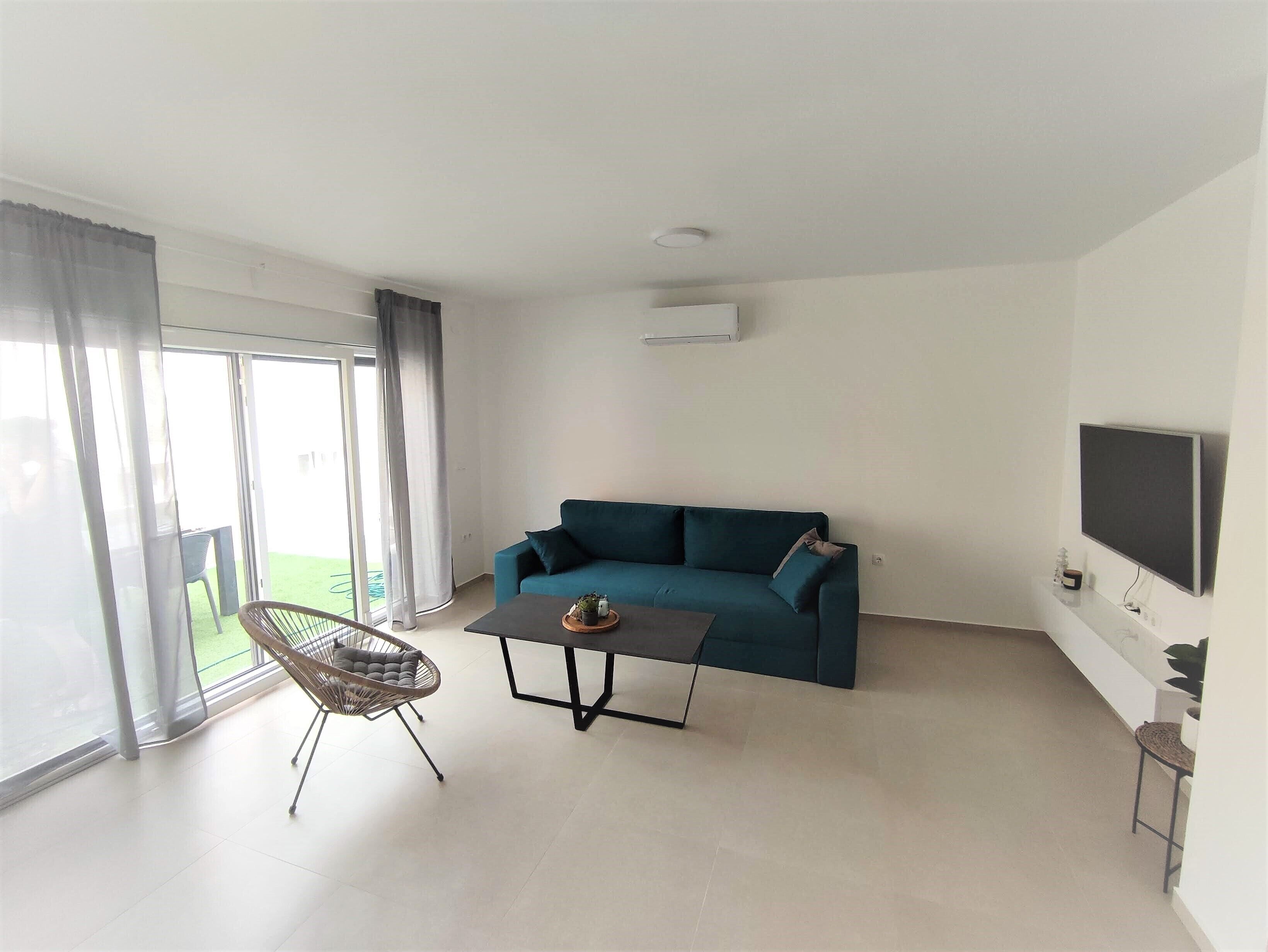 Una sala moderna con un sofá azul turquesa, una mesa de centro, una silla de mimbre y un televisor montado en la pared.