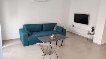 A modern living room with a teal sofa, a small coffee table, a wicker chair, a flat-screen TV mounted on the wall, and a potted plant on a side table.