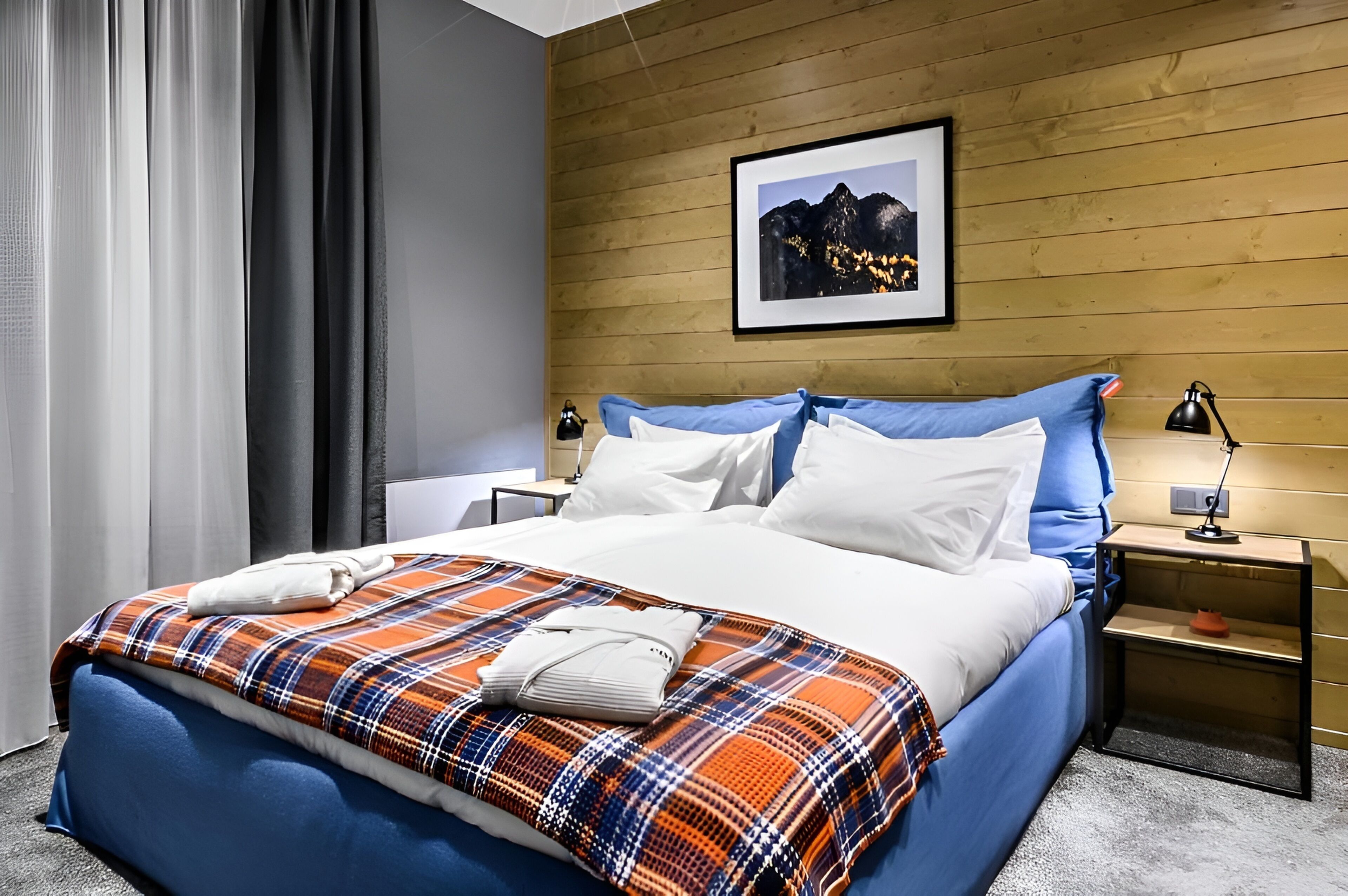 A hotel room with a bed, a nightstand, a lamp, a framed picture on the wall, and a window with curtains.