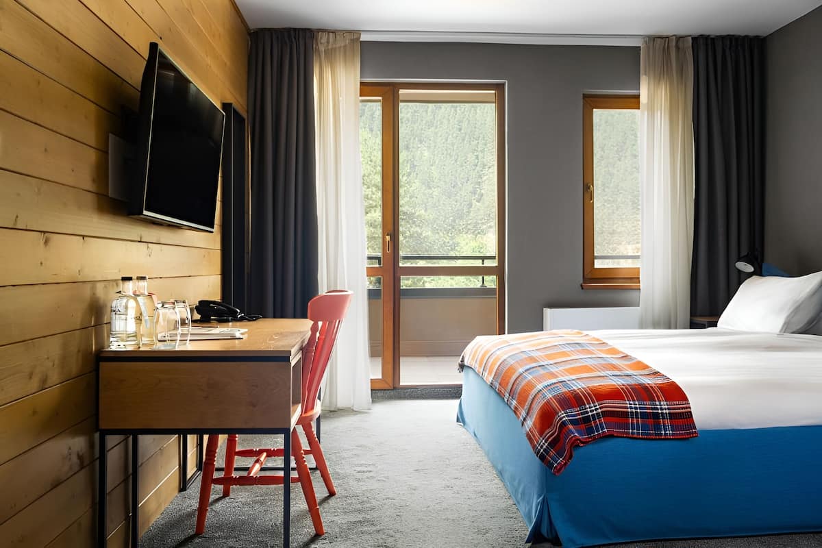 A hotel room with a wooden desk, a bed with a plaid blanket, and a TV mounted on the wall.