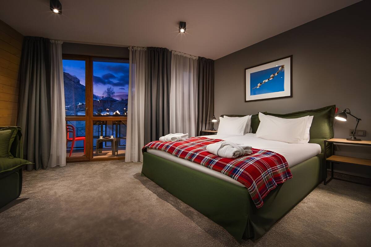 A hotel room with a large bed, a nightstand, a chair, a desk, and a view of the mountains through the window.