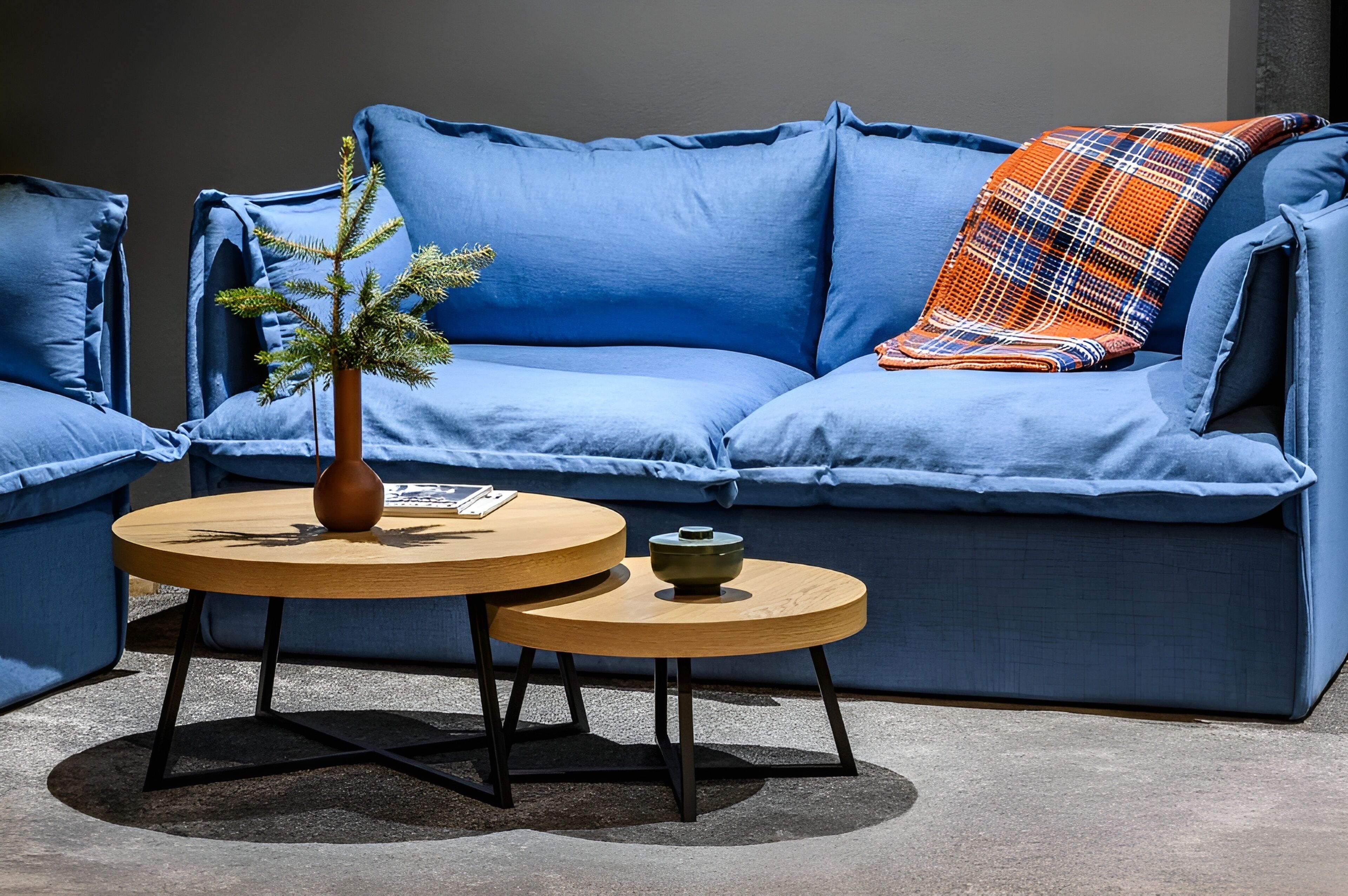 A blue sofa with a plaid throw pillow, two round wooden tables, and a small potted plant.