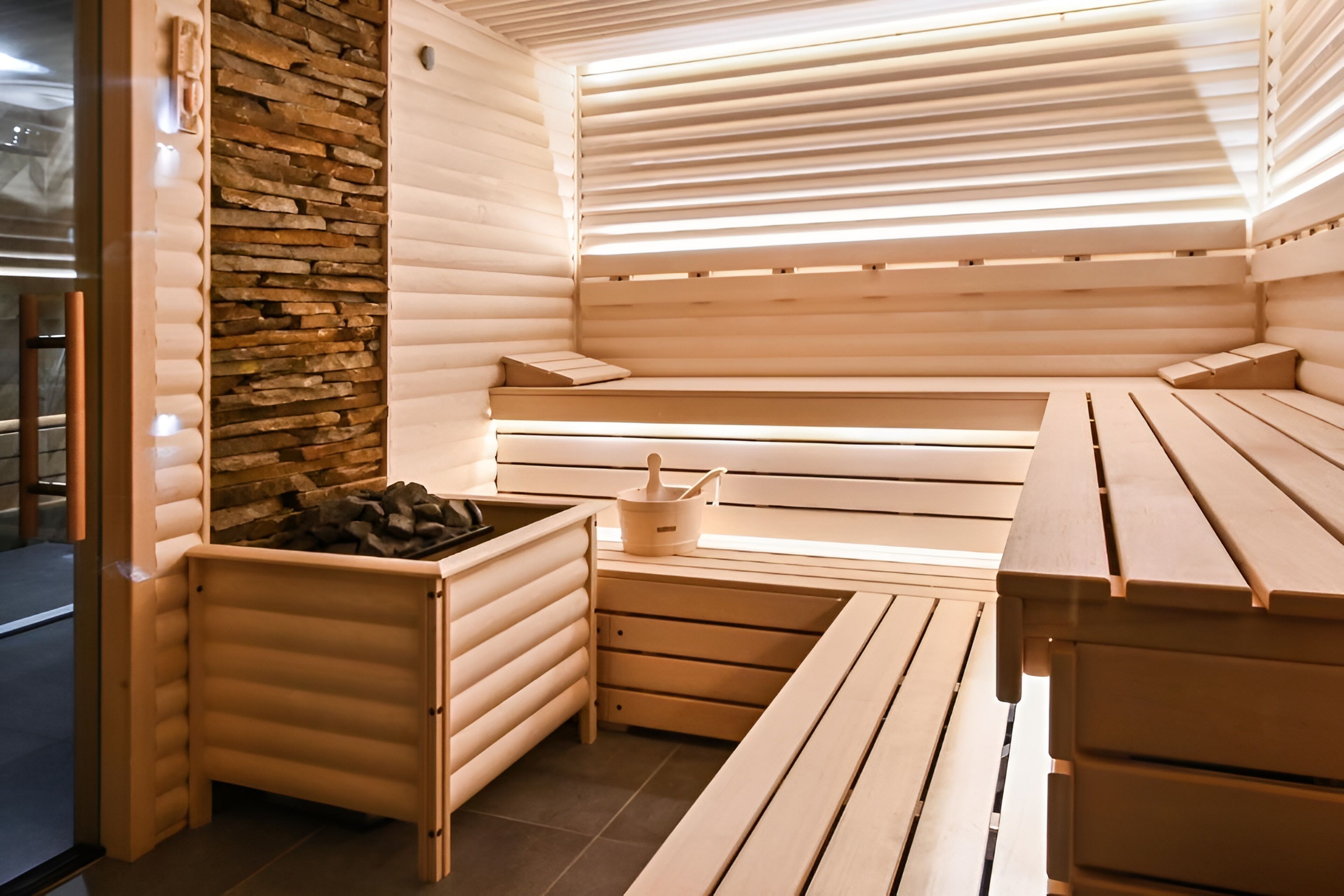 Una sauna con bancos de madera y una pared de piedra, que incluye un banco con una pequeña mesa y un cubo.