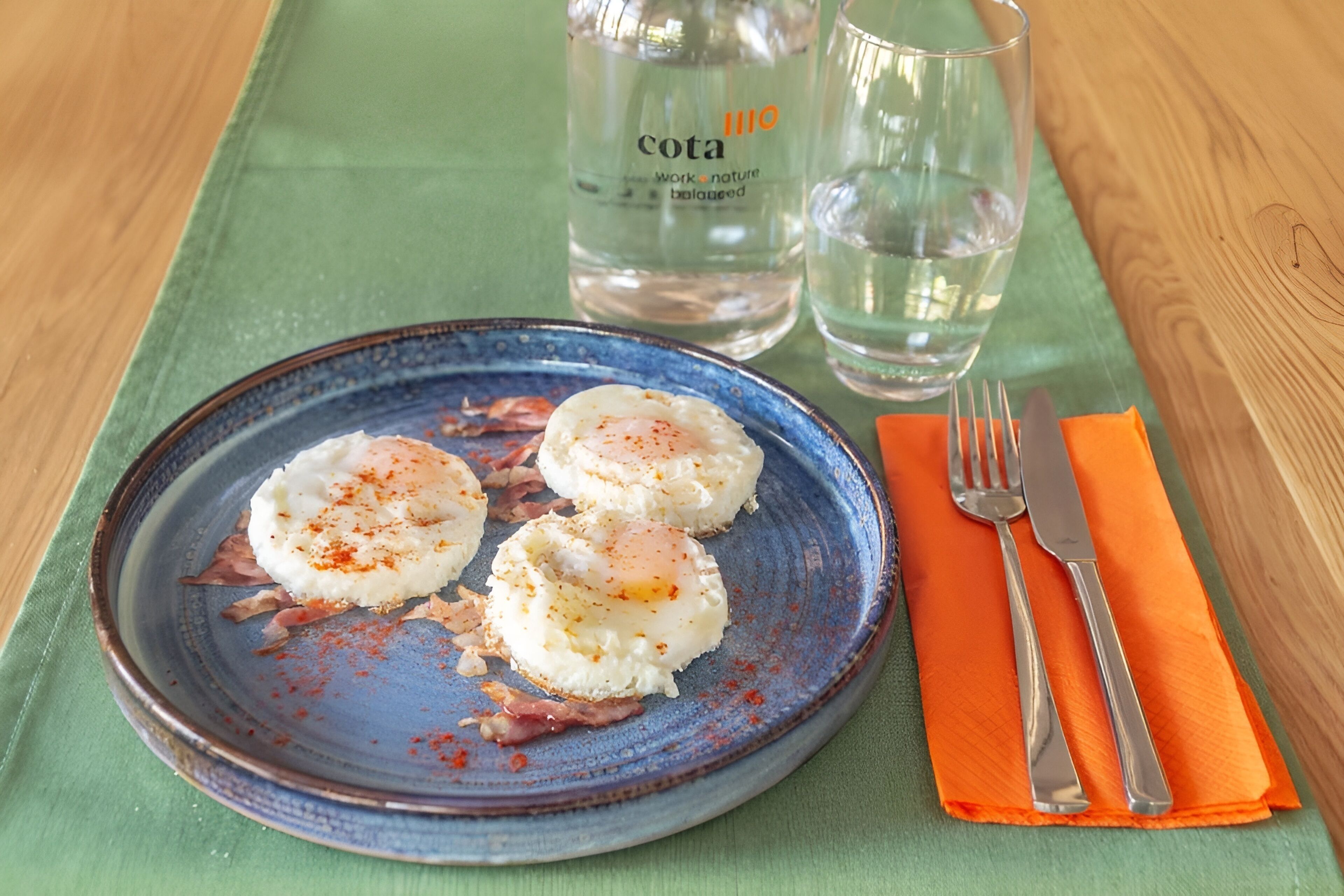 Un plato de comida con un borde azul, acompañado de un vaso de agua y una botella con la etiqueta 'Cota' sobre un mantel verde.