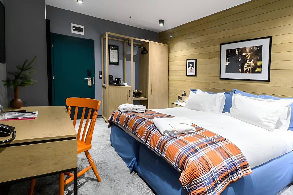 A hotel room with a bed, a desk, a chair, a wardrobe, and a small table with a plant.