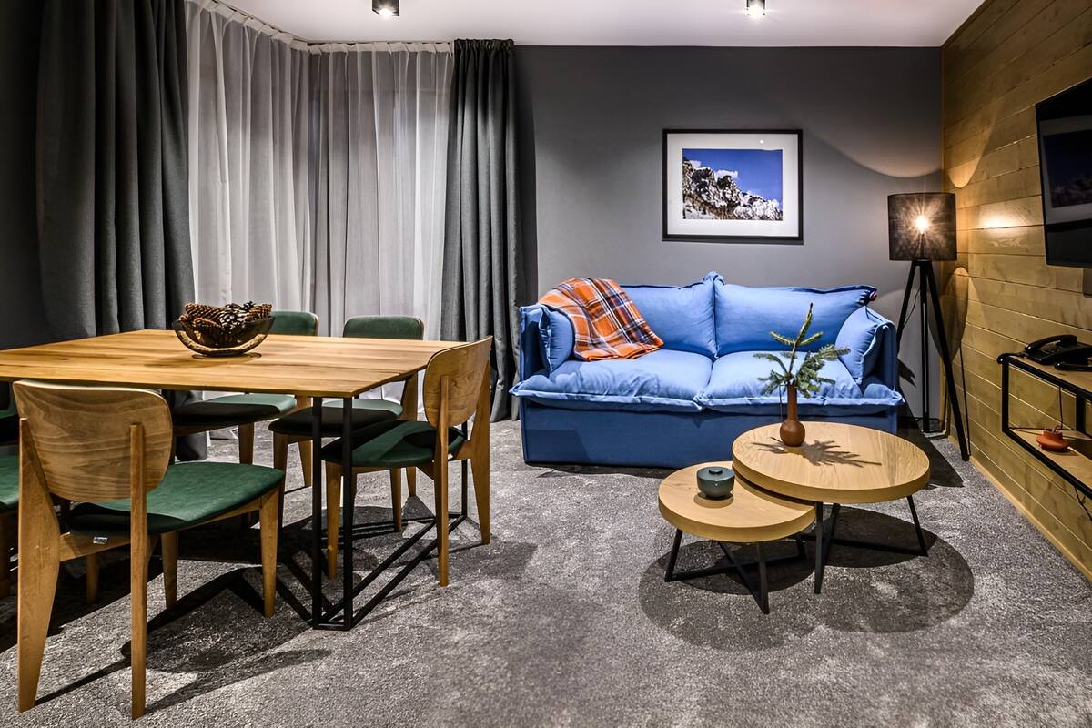 A modern living room with a wooden dining table, green chairs, a blue sofa with an orange throw, and a small round table with a plant.