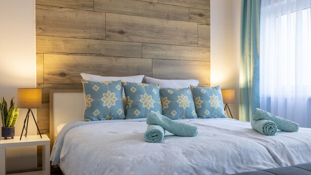 A neatly made bed with patterned pillows and rolled towels, a bedside plant, and a modern headboard.