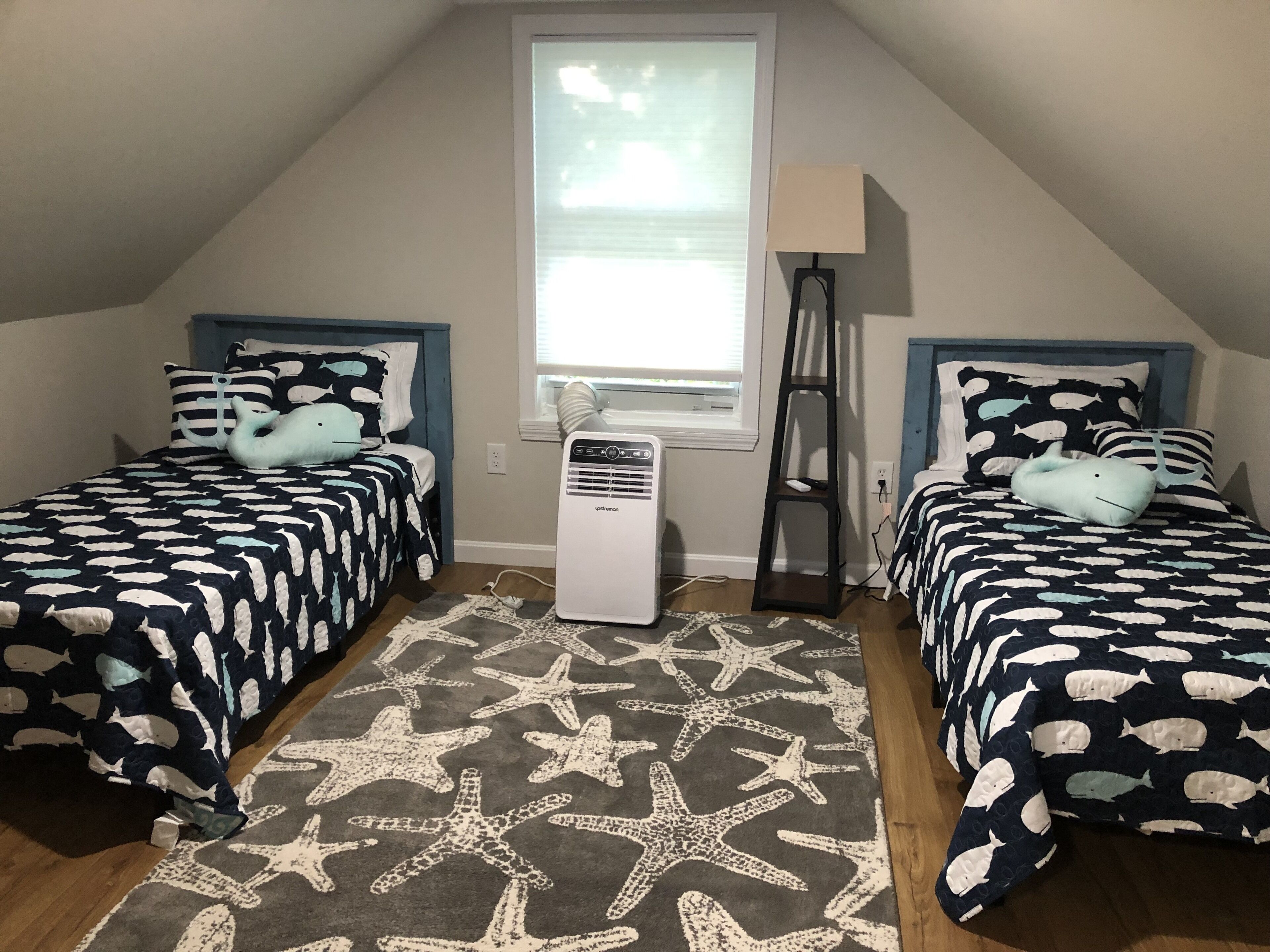A room with two beds, each with a blue headboard, a window with blinds, a standing lamp, and a starfish-patterned rug.
