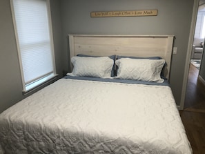 A bedroom with a bed, a window with blinds, and a motivational sign on the wall.