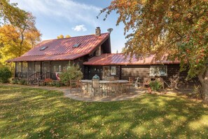 Un chalet rustique en rondins de bois, doté d’un toit en métal, d’un patio couvert avec des meubles d’extérieur et d’un jardin bien entretenu.