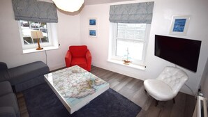 Una sala de estar con sofá, una silla roja, una mesa con mapa, televisor y ventana con persianas.