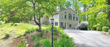 A two-story house with a front porch, surrounded by lush greenery and a driveway.