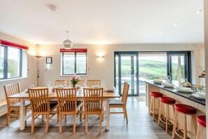 A dining area with a table set for four, wooden chairs, a vase of flowers, and a view through a glass door to the outdoors. - 6 Bed in Woolacombe (oc-mouro) (Woolacombe)