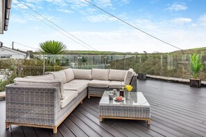 A modern outdoor seating area with a wicker sofa and a glass-top coffee table, overlooking a scenic landscape. - 6 Bed in Woolacombe (oc-mouro) (Woolacombe)