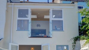 A two-story building with a balcony, a door leading to a room with a bed, and a small outdoor area with a plant.