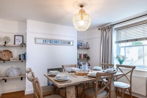 A dining area with a wooden table, chairs, a hanging light fixture, and shelves with books and decorative items.