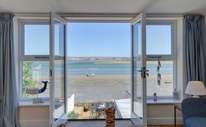 Een kamer met grote ramen met uitzicht op een strand, een klein tafeltje met stoelen en een lamp op een vensterbank.