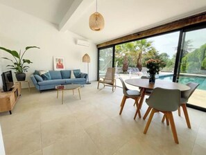 Een moderne woonkamer met een blauwe bank, een salontafel en een eethoek met een tafel en stoelen. Er is een groot raam met uitzicht op een zwembad en groen.