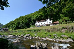 A riverside view of a white building with multiple windows and a red roof, surrounded by lush greenery and a stone wall.