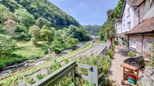 A riverside property with a stone building, a patio with outdoor furniture, and a lush green landscape. - 4 Bed in Lynmouth (oc-marins) (Barbrook)