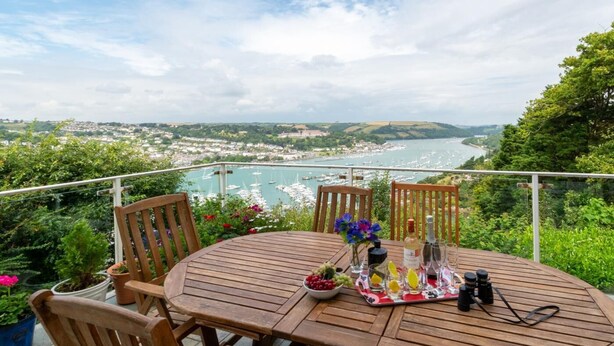 Une table en bois avec des chaises, une bouteille, des verres à vin et un appareil photo sur un balcon donnant sur un port, avec des bateaux et une ville au loin.