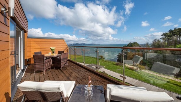 Une terrasse en bois, meublée pour l’extérieur, dotée d’une balustrade en verre et offrant une vue sur la mer et les collines.