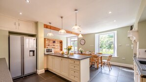 Cozinha com geladeira, micro-ondas, prateleiras de madeira, luminárias pendentes, área de refeições com cadeiras e uma janela com vista.