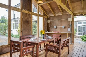Una estructura oberta de fusta amb taula de menjador preparada per a quatre, envoltada de cadires, i una zona de barbacoa amb vistes a una casa i un jardí.