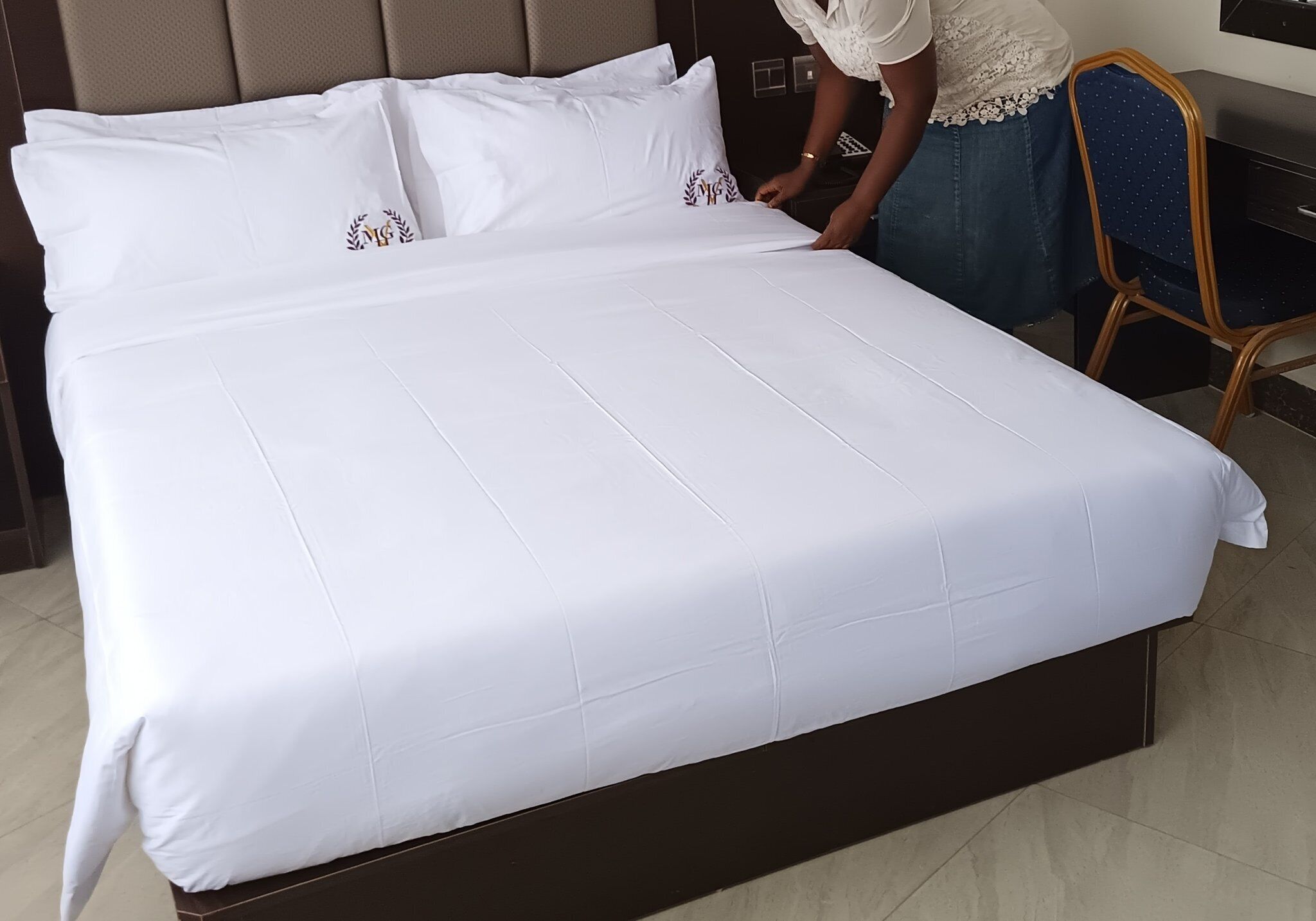 Een net opgemaakt bed met witte lakens en decoratieve kussens, een houten hoofdbord en een stoel met een blauwe hoes.