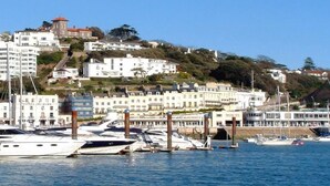 A marina with boats docked, a hotel named "Hotel Riviera" on the left, and buildings on a hillside in the background.