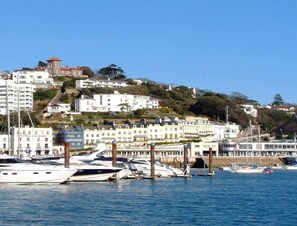 A marina with boats docked, a hotel named "Hotel Riviera" on the left, and buildings on a hillside in the background.