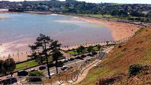 A coastal beach with a road and parking area in the foreground, a clear sky, and a distant town.