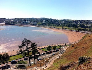 A coastal beach with a road and parking area in the foreground, a clear sky, and a distant town.