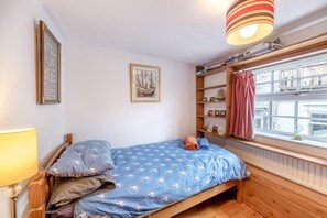 A small, cozy bedroom with a single bed, a window with a view, a shelf with books and a ship model, and a lamp on the left.