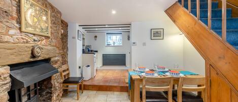 Un comedor acogedor con una mesa de madera dispuesta para cuatro personas, una chimenea de piedra y una escalera que lleva a un nivel superior.