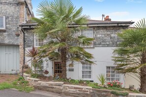 A two-story stone building with a white facade, a wooden door, and a small garden with palm trees and flowers. - 3 Bed in Torquay (oc-h75059) (Torquay)