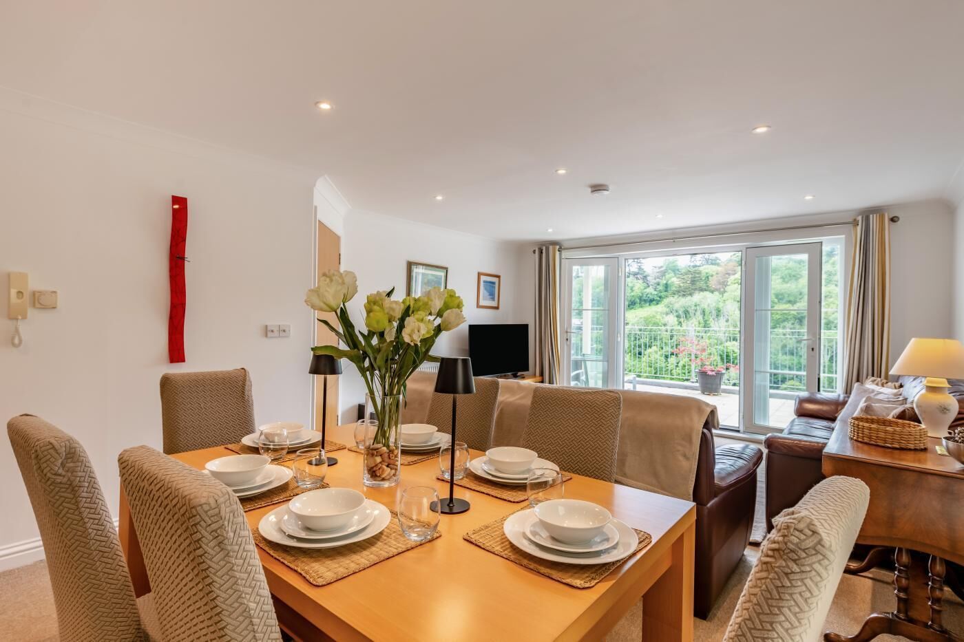 A dining area with a table set for four, chairs, a vase with flowers, and a television in the background.