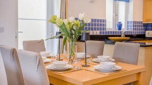 Uma área de refeições com uma mesa posta para quatro pessoas, um vaso de flores e vista para a cozinha ao fundo.